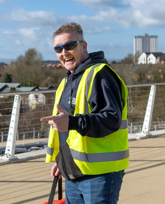 A lecturer in a Glasgow Kelvin College hoodie and high-visibility vest smiling and pointing towards the camera while carrying a bright orange equipment case on a bridge. A lecturer in a Glasgow Kelvin College hoodie and high-visibility vest smiling and pointing towards the camera while carrying a bright orange equipment case on a bridge.